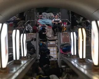 People shelter on a Kyiv metro platform during a missile strike, viewed through handrails, with luggage and blankets