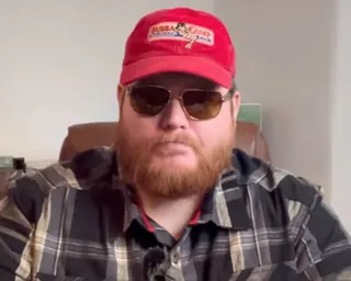 Bonehill-Paine, bearded and wearing sunglasses and a red cap, sits in a plaid shirt