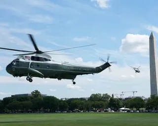 Helicopter flying in sky over grassy lawn with a building, trees and another helicopter in the background