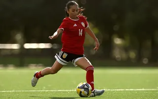 Action shot of a woman about to kick a football. She is wearing a red shirt with the number 14 on it in white. She has black shorts, red socks and white boots. Her long dark hair is tied in a ponytail.