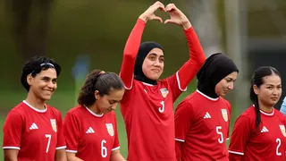 Five women in red football kit stand in a line. The one in the middle is holding her arms above her head and her hands form the shape of a heart.