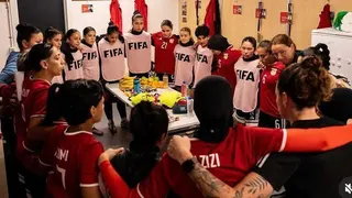 About 20 women huddle in a circle in a changing room with their arms around each other. Some wear red football shirts and some have white bibs over their kit with the word Fifa written in dark blue. There is a table in the middle of the circle with a few bottles and some fruit on it.