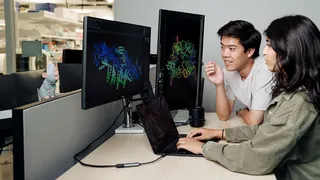 Two people working together at a desk in what appears to be a laboratory or research office environment. Large computer monitors display colorful three-dimensional molecular or protein structure models against dark backgrounds, while a laptop on the desk shows lines of code or data. One person is seated using the laptop, and another is leaning toward the screens as both focus on the scientific visualizations. Laboratory shelving and equipment are visible in the background, suggesting a biomedica