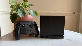 Steam Machine photograph showing the steam machine with black faceplate on a marble circular table in front of a grey/beige wall, with a plant pot and the Steam Controller next to it