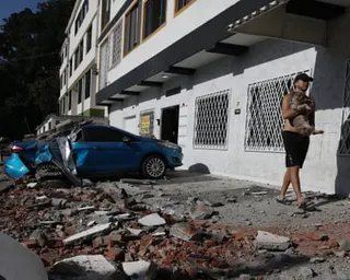 A man carries his dog past a car crushed by rubble