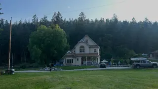 An image showing Keegan Radomski's home - a two-storey house with a slanted roof and an attic, a drive way with two cars visible parked nearby. Behind it is a row of large spruce trees. In front of it is a large grass field.