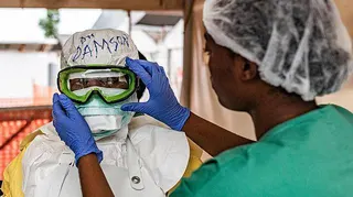 A person in PPE and full head covering is having their goggles adjusted by someone, with blue protective gloves, out-of-focus in the foreground. The green scrubs and hair covering of that person can be seen on the right of the picture.