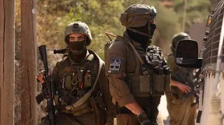 Two IDF soldiers in military gear and face coverings block a road near the West Bank village of Qusra near Nablus. One soldier is seen facing towards the side of a van while a second holding a rifle is facing towards the camera but looking off in the distance.