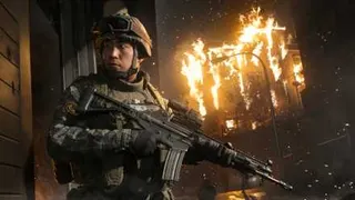 A man in modern military regalia with an assault rifle in front of a burning building.