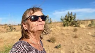 Eva Greztmacher looks up in sunglasses. A blue sky and desert scrubland can be seen behind her. She is wearing a necklace with a ring as a pendant.