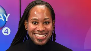 Prof Arday is smiling. He's wearing a black top and has earrings in. A backdrop with an ombre of colours is behind him.