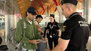 Two police officers with black uniforms look at documents of two people - one wearing a green hoodie and a blue baseball cap and carrying a grey backpack on his right shoulder, the other has a light T-shirt and is pulling something out of an apparent wallet