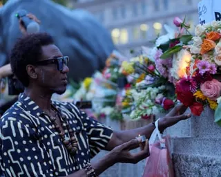 A man places flowers among a floral memorial outside a public building, with a statue and city buildings in the background.