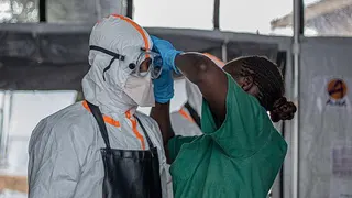 A health worker in green scrubs helps another person put on their PPE. The person on the left of the picture is wearing full white protective gear and goggles.