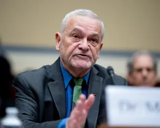 Man in suit gestures with hand as he sits at a table with a microphone