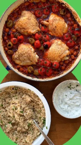 A pot of sauce seen from above with four chicken thighs nestled in it. A bowl of yoghurt dressing, and another bowl containing rice are standing next to it.