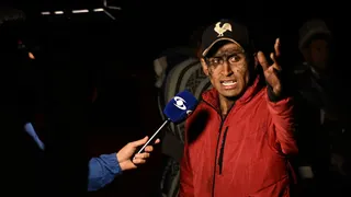 The relative of a miner caught in the explosion at the mine in Sutatausa gestures as he speaks to the media. His face is stained with coal dust. He is wearing a black baseball cap with a white cockerel embroidered on top and a red coat.
