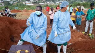 Two people in blue PPE stand over a coffin as they lower it into the ground. Some young boys can be seen in the background looking on.