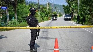 A soldier completely clad in a black uniform and holding a gun stands at a yellow police tape which stretches over a road. A red traffic cone is in the foreground.