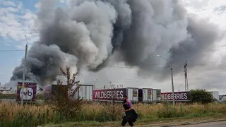 Wildberries employee walks past plumes of smoke rising above wildberries logistics  complex. There are seven company lorries in front of the smoke as well as roadside shrubs