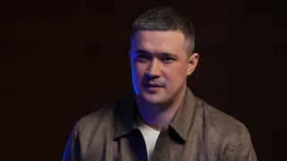 A man dressed in a white T-shirt and brown collared shirt in front of a black background sits talking to someone with their back to the camera.