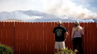 Two people look over what appears to be a domestic fence. They are looking at where smoke is rising from hills in the background.