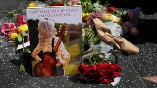 2026年8月25日，在美國加州洛杉磯，鄉村音樂傳奇人物 Dolly Parton 逝世，終年80歲的消息公布後，人們在她位於荷里活星光大道上的星形紀念章前獻上鮮花。花束左側豎立了一張  Dolly Parton 背著吉他，背對鏡頭，望向陽光的照片，照片頂部印上字句：「告別廣受愛戴的美國傳奇。」