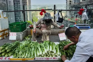 2025年4月17日，中國重慶，兩名郊區農民在高架人行道上的非正式攤位上販售新鮮採摘的蔬菜，男子正在整理農產品，女子坐在他身後。