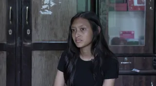 A girl with dark hair sits being interviewed in front of a door, she is in a black t-shirt.