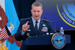 man in blue suit with military pins points while speaking at a podium marked 'department of war'