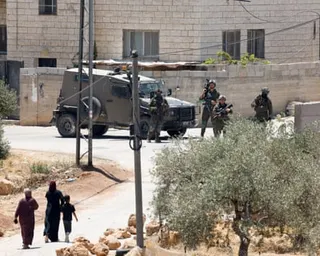 Two Palestinian women and a child walk up a road towards a junction guarded by armed troops and a vehicle