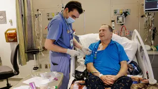 The image shows a hospital room with a patient seated in an adjustable bed, wearing a bright blue polo shirt and patterned dark trousers. A healthcare worker dressed in blue scrubs, gloves, a face mask, and a stethoscope is standing beside the bed and handling tubing connected to a medical access point on the patient's upper chest. Various items including syringes, medical packaging, paperwork, and a disposable cup are arranged on a bedside table in the foreground. The room contains hospital equ
