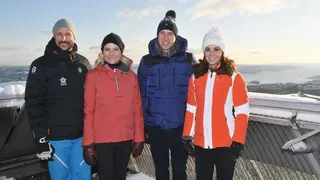 Four people stand smiling at the camera in ski jackets and woollen hats. On the left is a man with a goattee beard and next to him is a woman with blonde hair. Next is Prince William wearing a blue ski jacket and pom-pom hat, and Kate wearing a bright orange jacket and white hat. They are standing outside and look to be high-up on a mountain, and in the background is snow and a view.