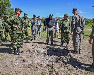 A man in a cap surrounded by police and military personnel looking at a hole in the ground