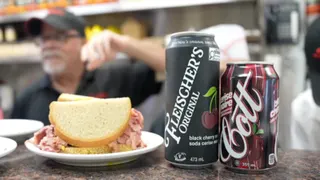 A sandwich and two types of cherry cola on a countertop in a deli.