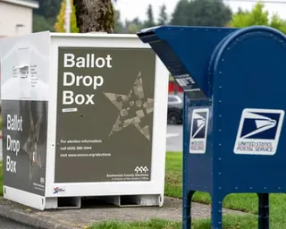 A white ballot drop box stands beside a blue USPS mail collection box in Bothell, Washington