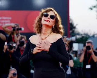 Susan Sarandon poses in sunglasses and an off-shoulder black dress as photographers shoot behind her