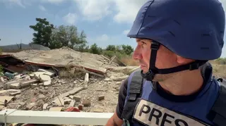BBC correspondent Hugo Bachega, wearing a blue flak jacket and helmet, sits on top of a Unifil vehicle as it drives past destroyed buildings in southern Lebanon